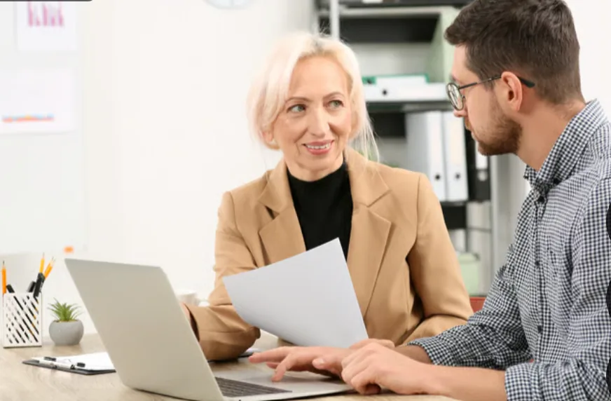 Two colleagues at an office desk going through a printed document together at a laptop.